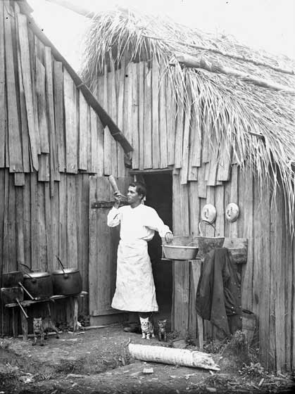 The cook blows on a bullock horn to call the bushmen to a meal - bullock horns were also used to raise the alarm if there was an accident in the bush. The structure to the left of the main hut is a huge chimney containing the cooking fire and equipment. The rough slab hut has a thatched roof, probably made of nikau leaves. Photo